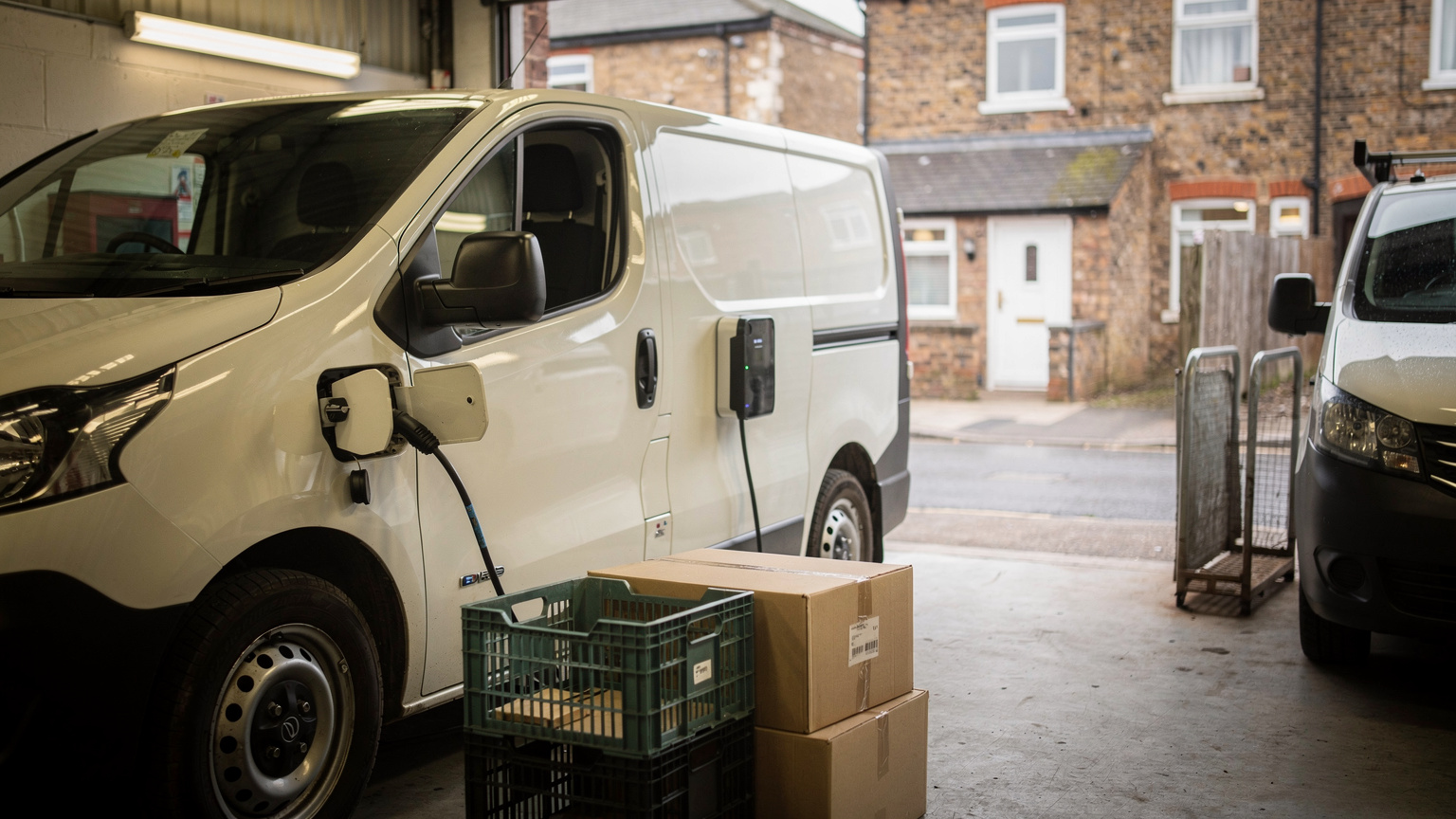 A white electric van plugged into a charging point inside a depot loading bay, with crates and cardboard boxes stacked beside it and a second van nearby.