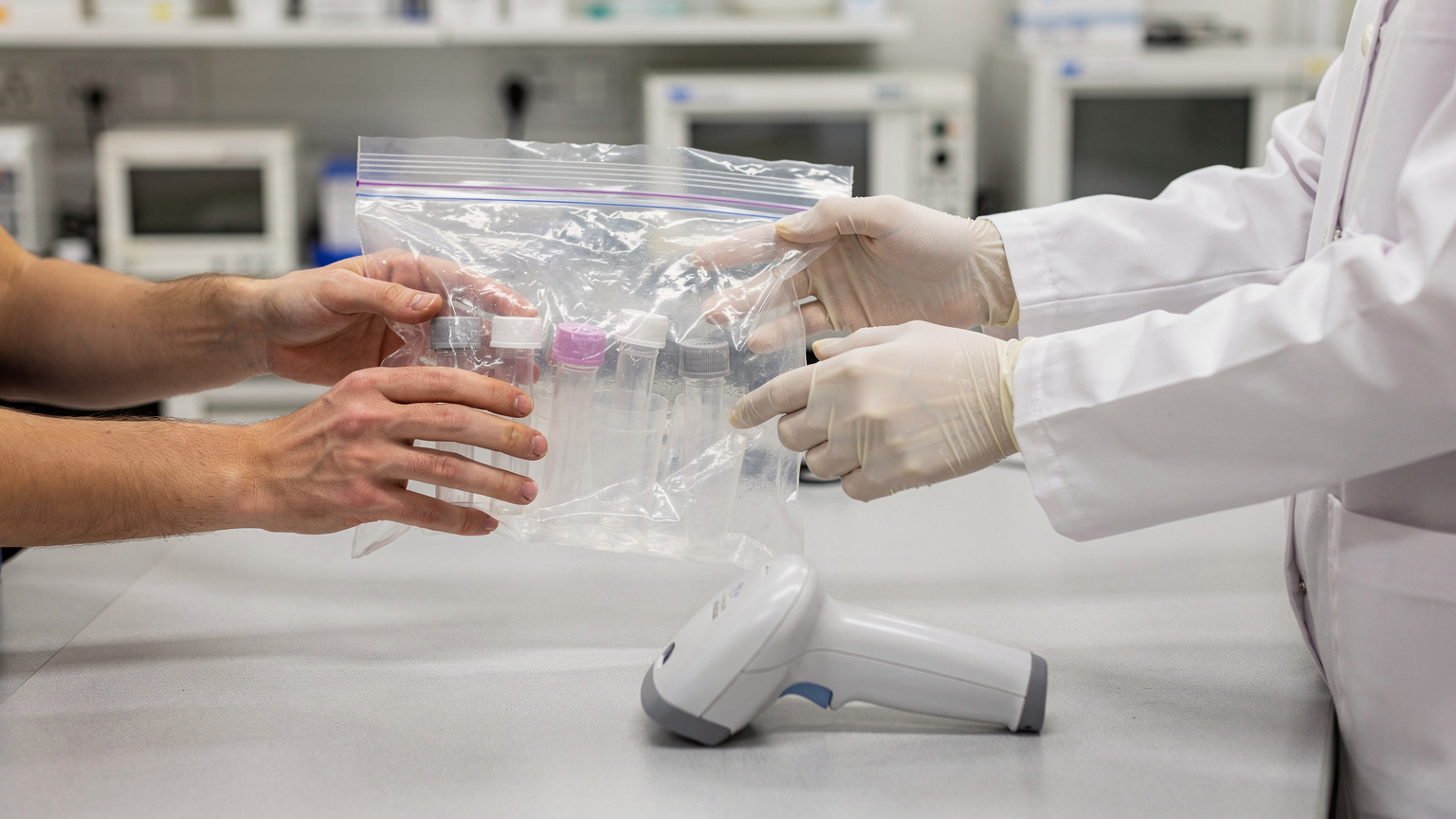 A courier's hands pass a sealed clear specimen bag of sample tubes to a gloved lab technician, a handheld scanner resting on the counter.